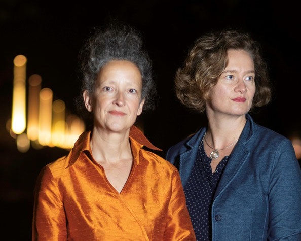 AI generated: The image shows two women looking into the distance and standing in front of a dark background with blurred lights. One woman is wearing an orange-coloured top, while the other is wearing a blue blazer.