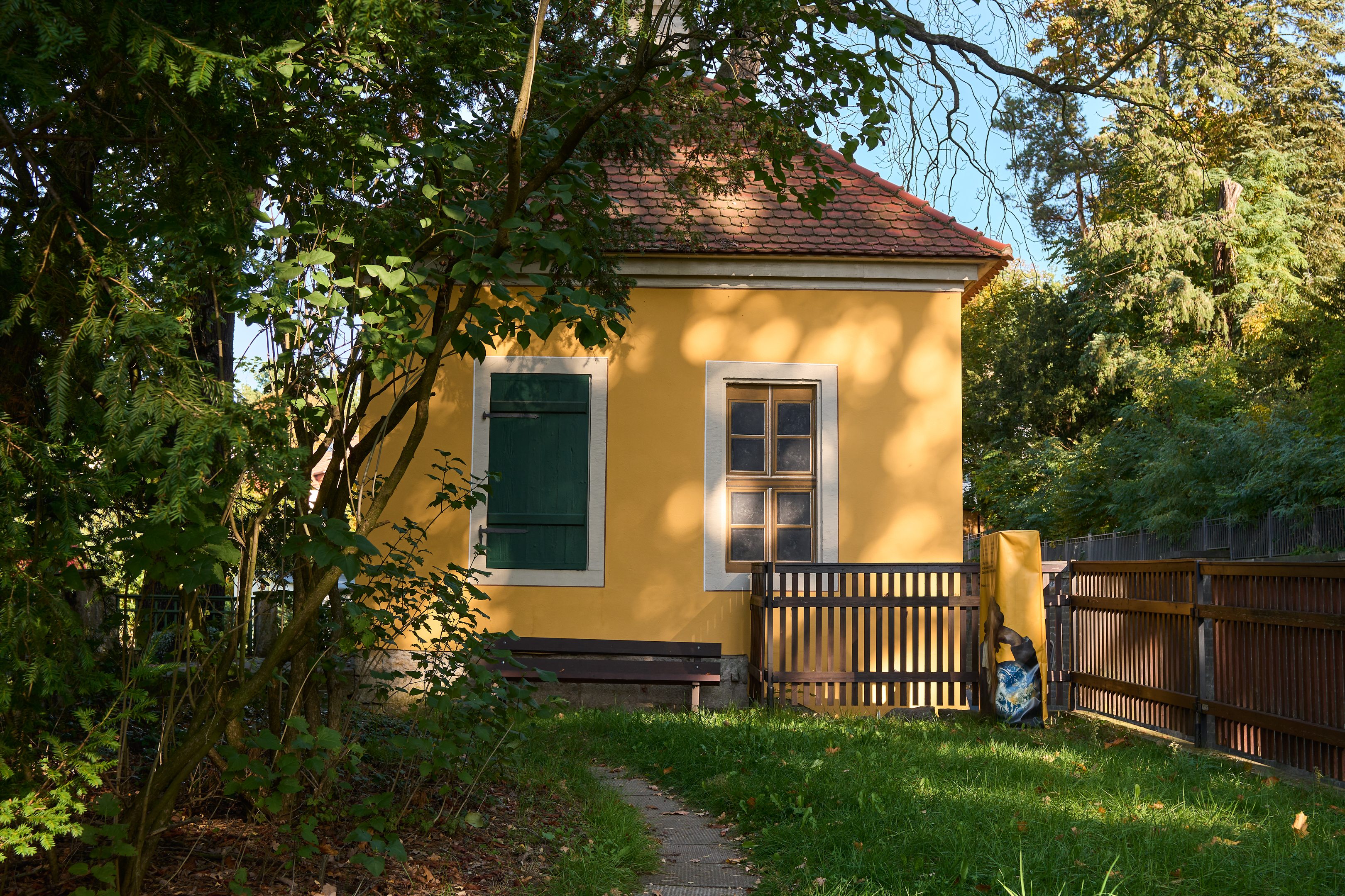 KI generiert: Das Bild zeigt ein kleines gelbes Haus mit roten Ziegeldach und grünen Fensterläden, umgeben von Bäumen und einem Garten. Das Haus steht hinter einem Holzzaun, und auf der linken Seite befindet sich eine Bank.
