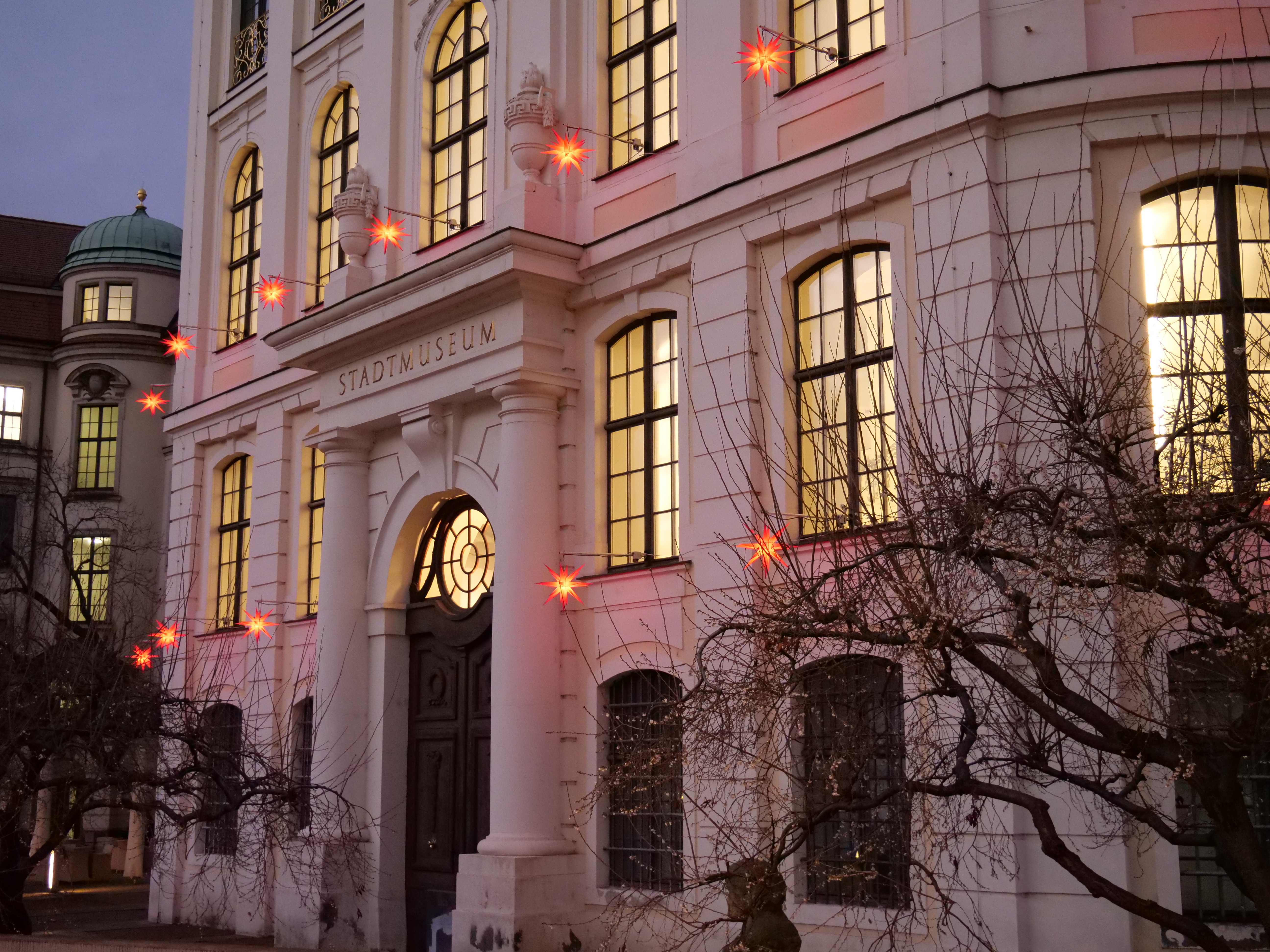 KI generiert: Das Bild zeigt die Fassade eines Stadtmuseums, dekoriert mit leuchtenden Sternen und erleuchteten Fenstern. Im Vordergrund sind kahle Bäume zu sehen.