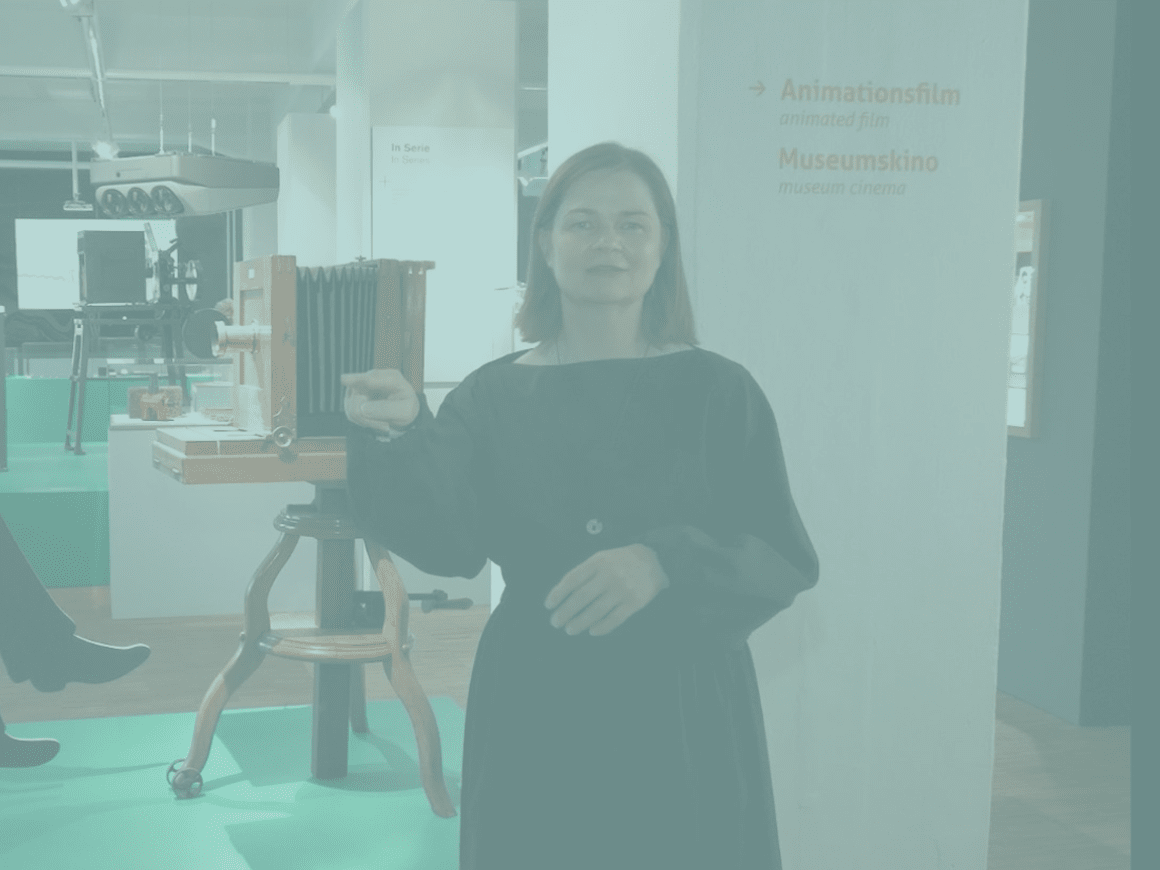 KI generiert: Das Bild zeigt eine Frau in einem Museum, die vor einer antiken Kamera steht. Im Hintergrund sind weitere Ausstellungstücke zu sehen, sowie ein Schild, das auf das Animationsfilm- und Museumskino hinweist.