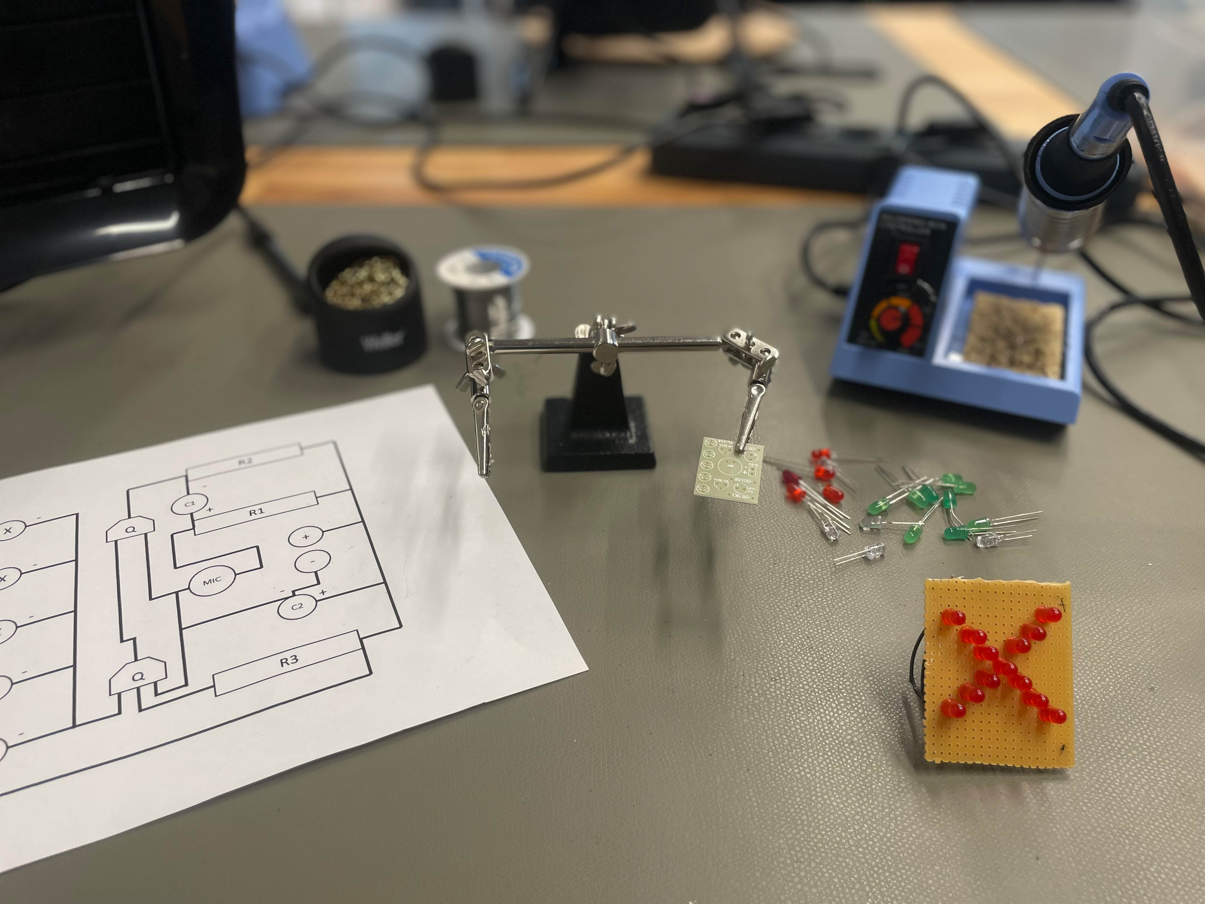 KI generiert: Auf dem Bild sind elektronische Komponenten und Werkzeuge auf einem Tisch zu sehen, darunter eine "dritte Hand" mit einer Platine, ein Schaltplan sowie Lötutensilien und LEDs. Der Hauptinhalt scheint sich auf die Vorbereitung oder Durchführung eines Lötprojekts zu konzentrieren.