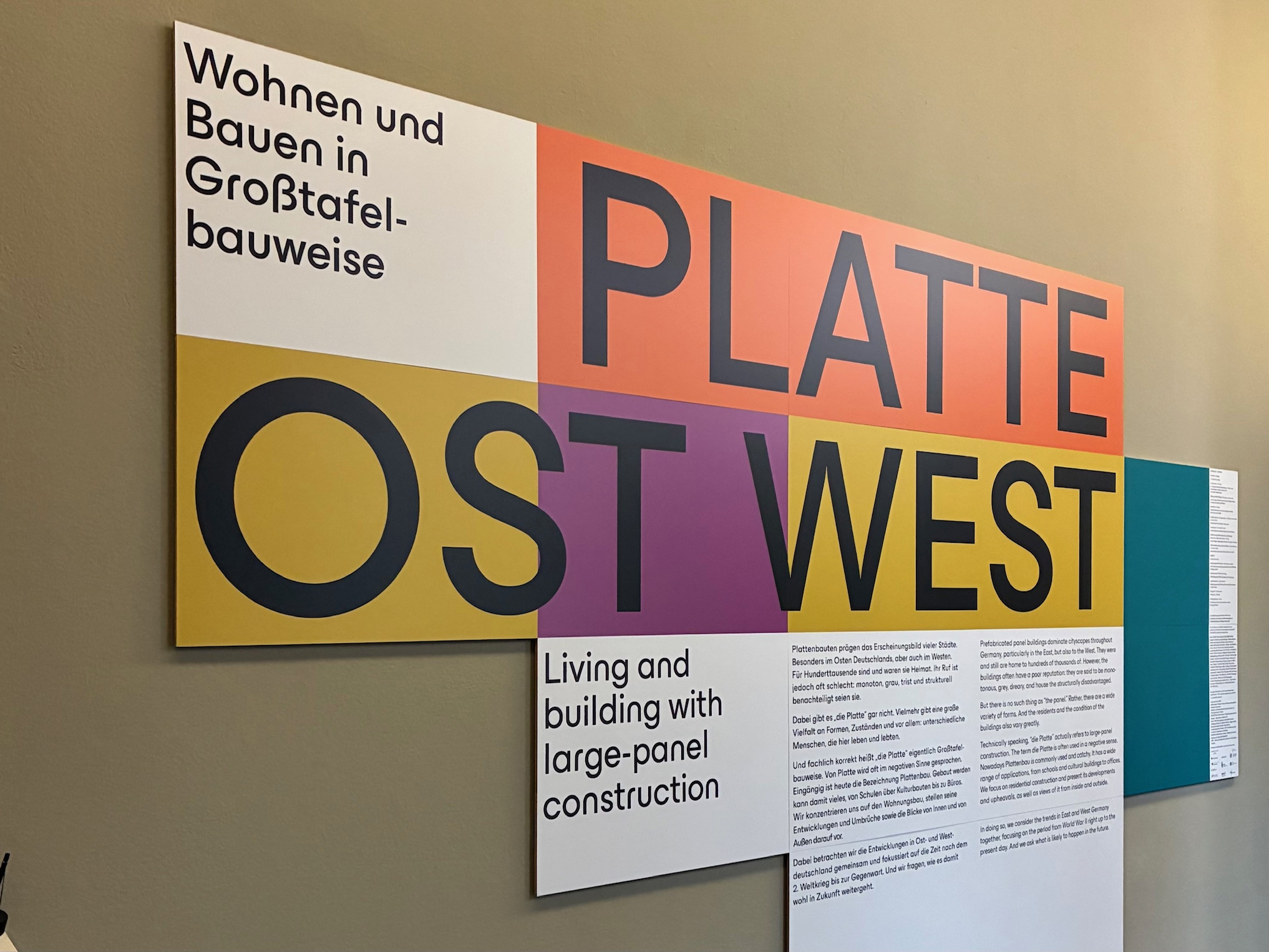 KI generiert: Das Bild zeigt eine bunte Infotafel mit der Überschrift "PLATTE OST WEST", die sich mit Großtafelbauweise im Wohnungsbau beschäftigt. Es gibt Erklärungstexte auf Deutsch und Englisch zu diesem Baukonzept.