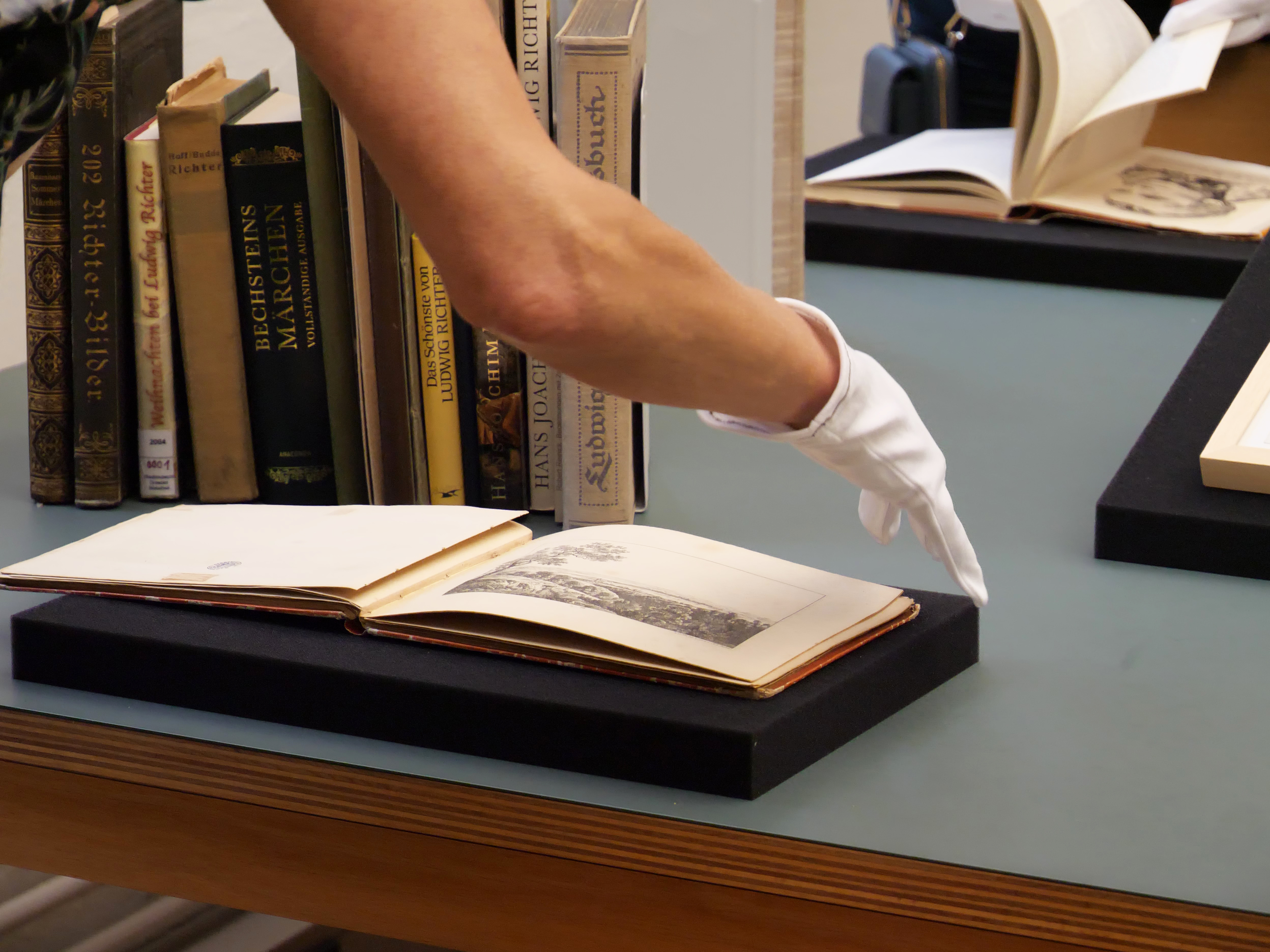 KI generiert: Das Bild zeigt eine Hand mit einem weißen Handschuh, die ein altes aufgeschlagenes Buch betrachtet, das auf einem Podest liegt. Im Hintergrund stehen mehrere Bücher in einem Regal.