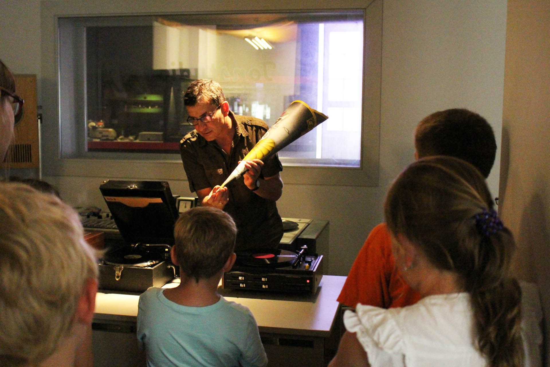 KI generiert: Ein Mann zeigt einer kleinen Gruppe von Kindern ein neues Musikinstrument oder ein altes Tonabspielgerät. Sie stehen alle um ihn herum und scheinen neugierig zuzuhören.