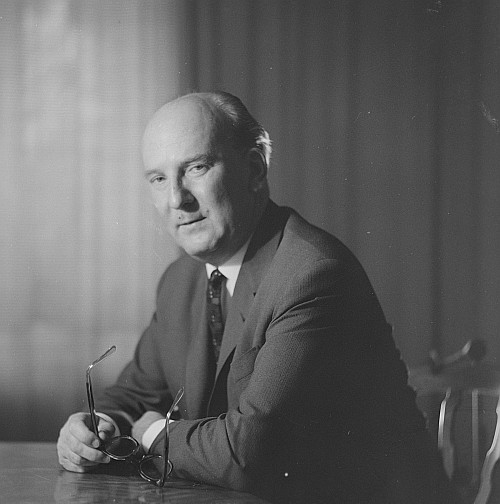 KI generiert: Das Bild zeigt einen Mann in formeller Kleidung, der an einem Tisch sitzt und eine Brille hält. Der Hintergrund wirkt neutral und schlicht.