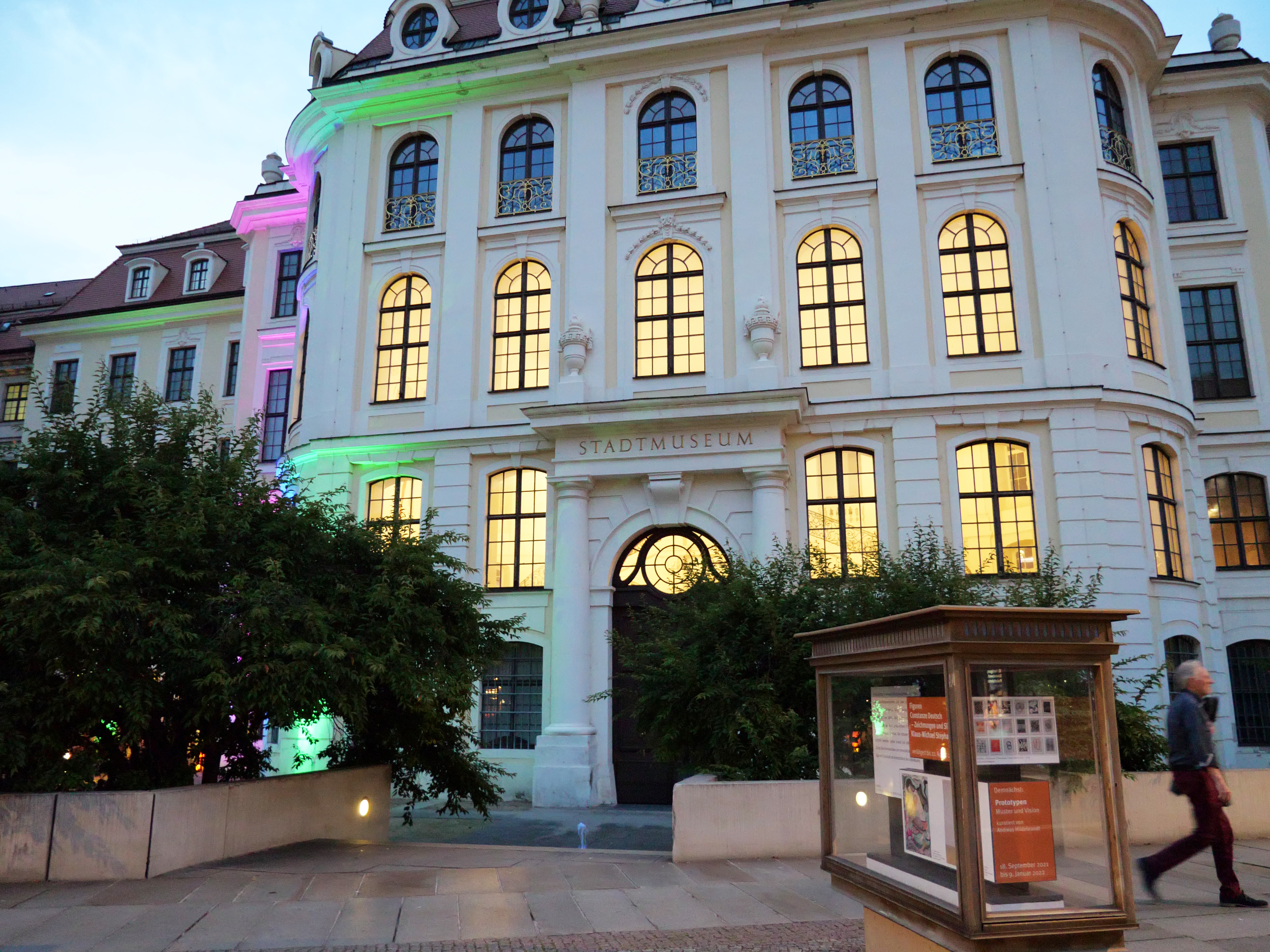 AI generated: The image shows the illuminated façade of a city museum with magnificent architecture and high windows. Trees and an information display case are visible in front of the building.