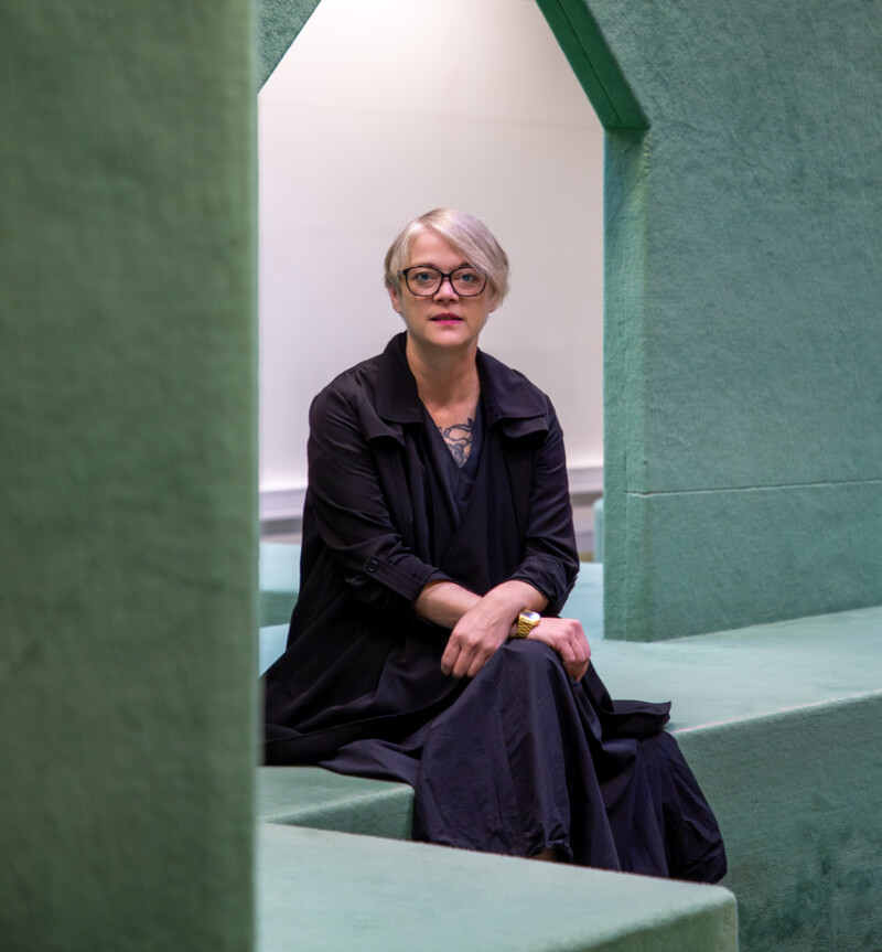 AI generated: The image shows a person with glasses and short, blonde hair sitting on a green, architecturally interesting structure. The surroundings look modern and minimalist.