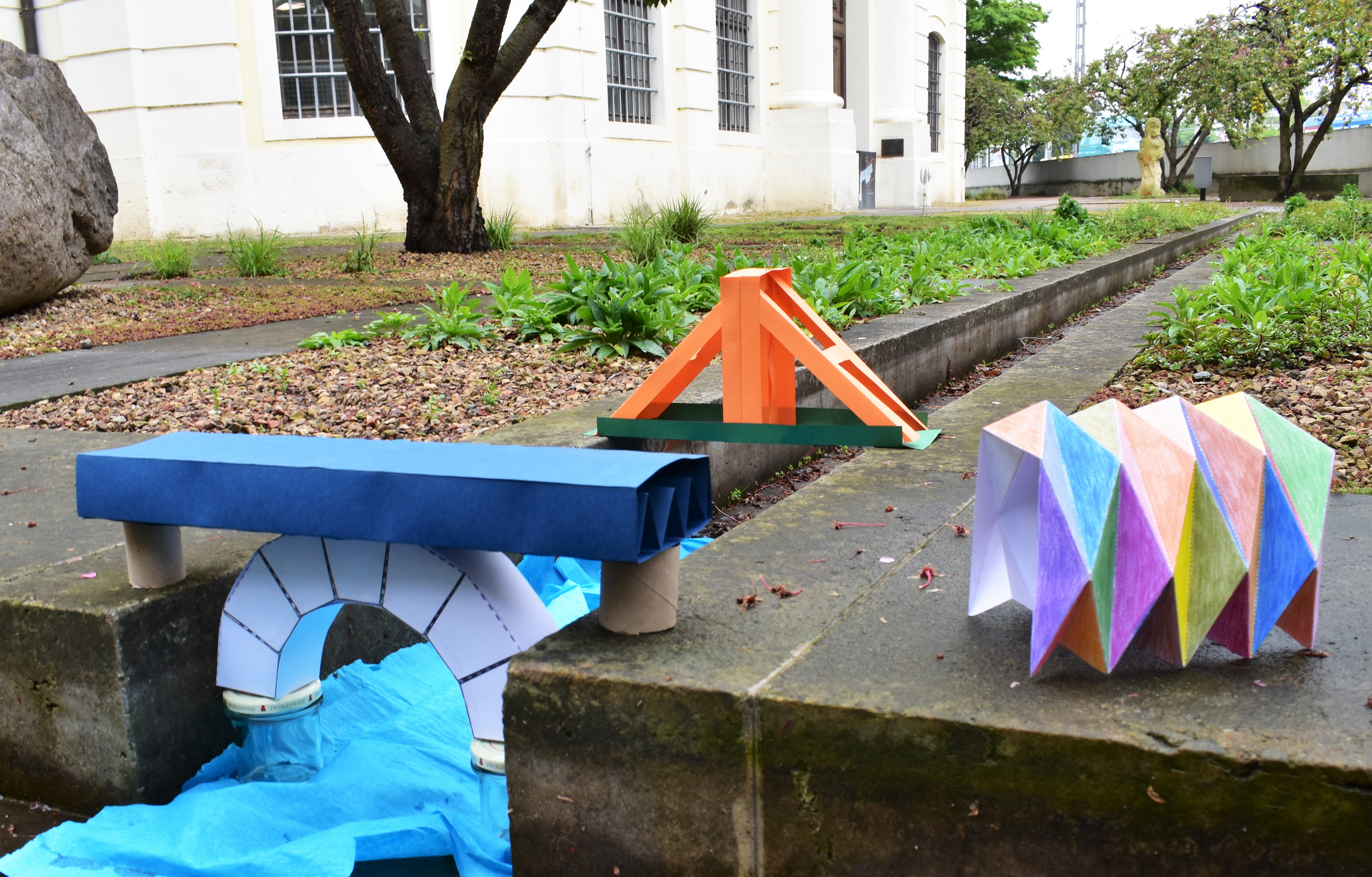 KI generiert: Das Bild zeigt verschiedene, aus Papier und Karton gebastelte Brückenmodelle, die auf einer Steinfläche im Freien stehen. Es handelt sich um eine kreative Darstellung mit einem Fokus auf Architektur und Design.