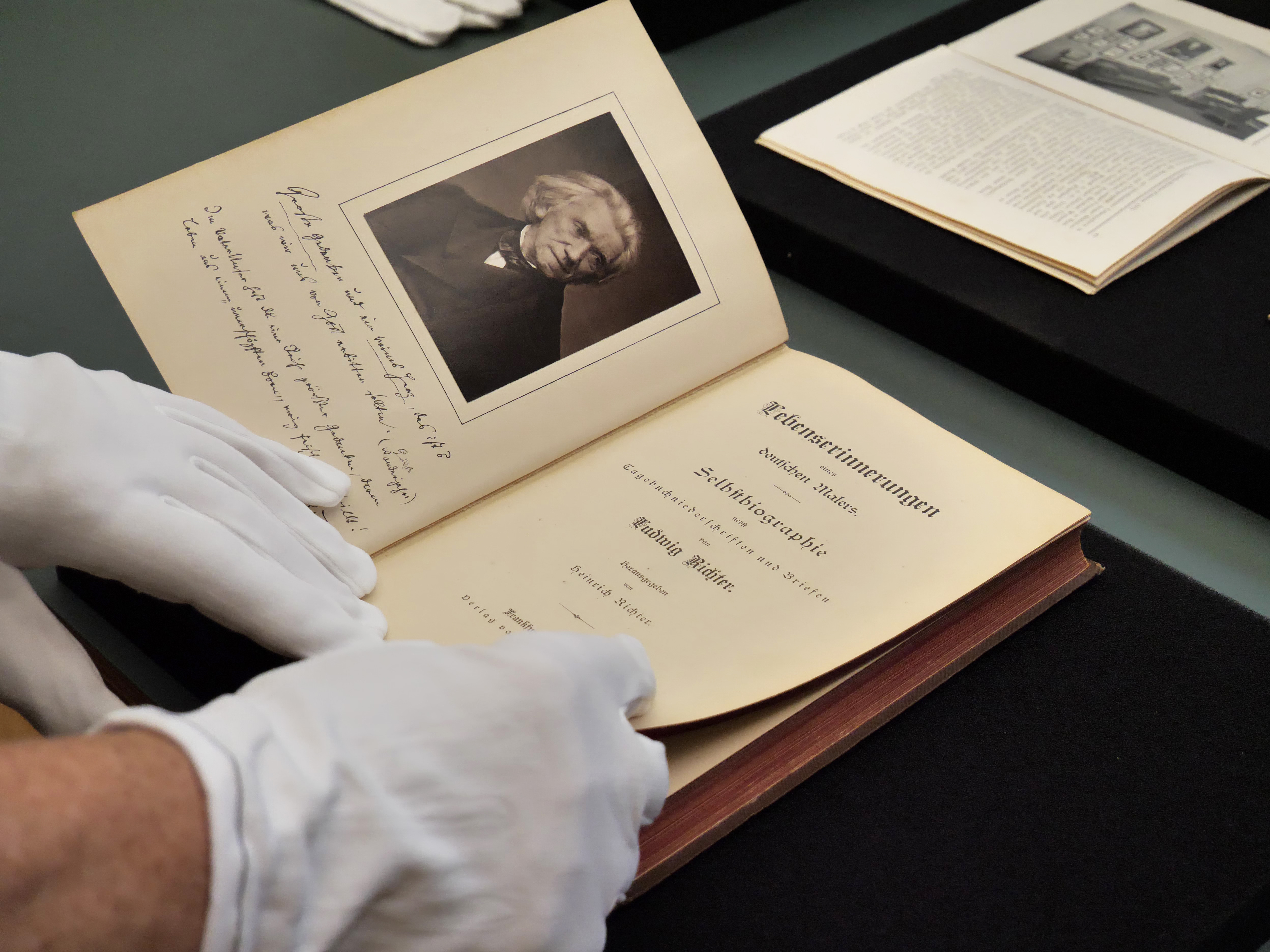 KI generiert: Das Bild zeigt Hände in weißen Handschuhen, die ein altes Buch öffnen, auf dessen Seiten ein Porträt und eine Seite mit Text zu sehen sind. Der Hauptinhalt ist die sorgfältige Handhabung eines historischen Dokuments oder Buches.