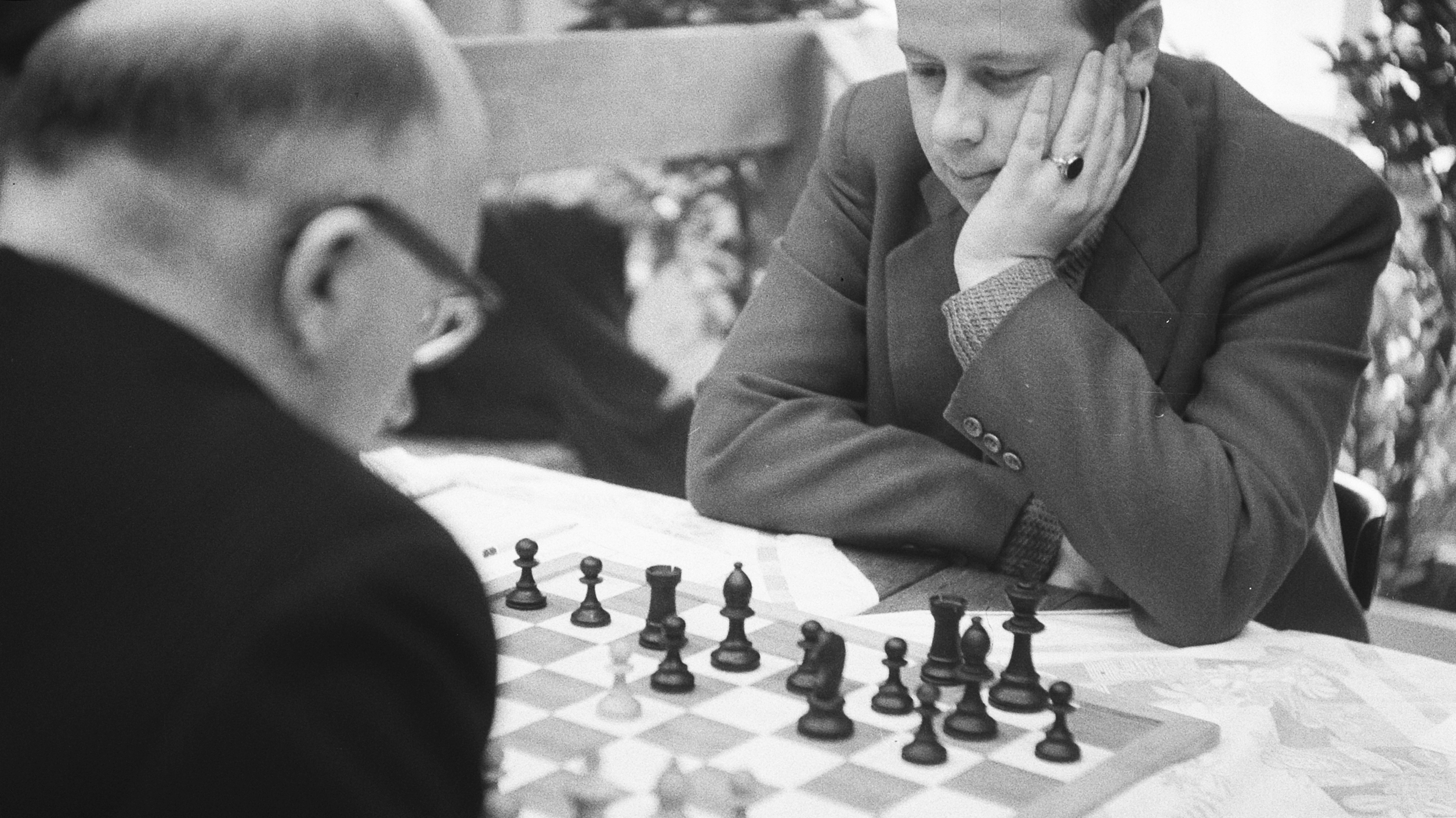 AI generated: The image shows two people playing a concentrated game of chess. One person can be seen in the foreground from behind, while the other is bent over the chessboard.