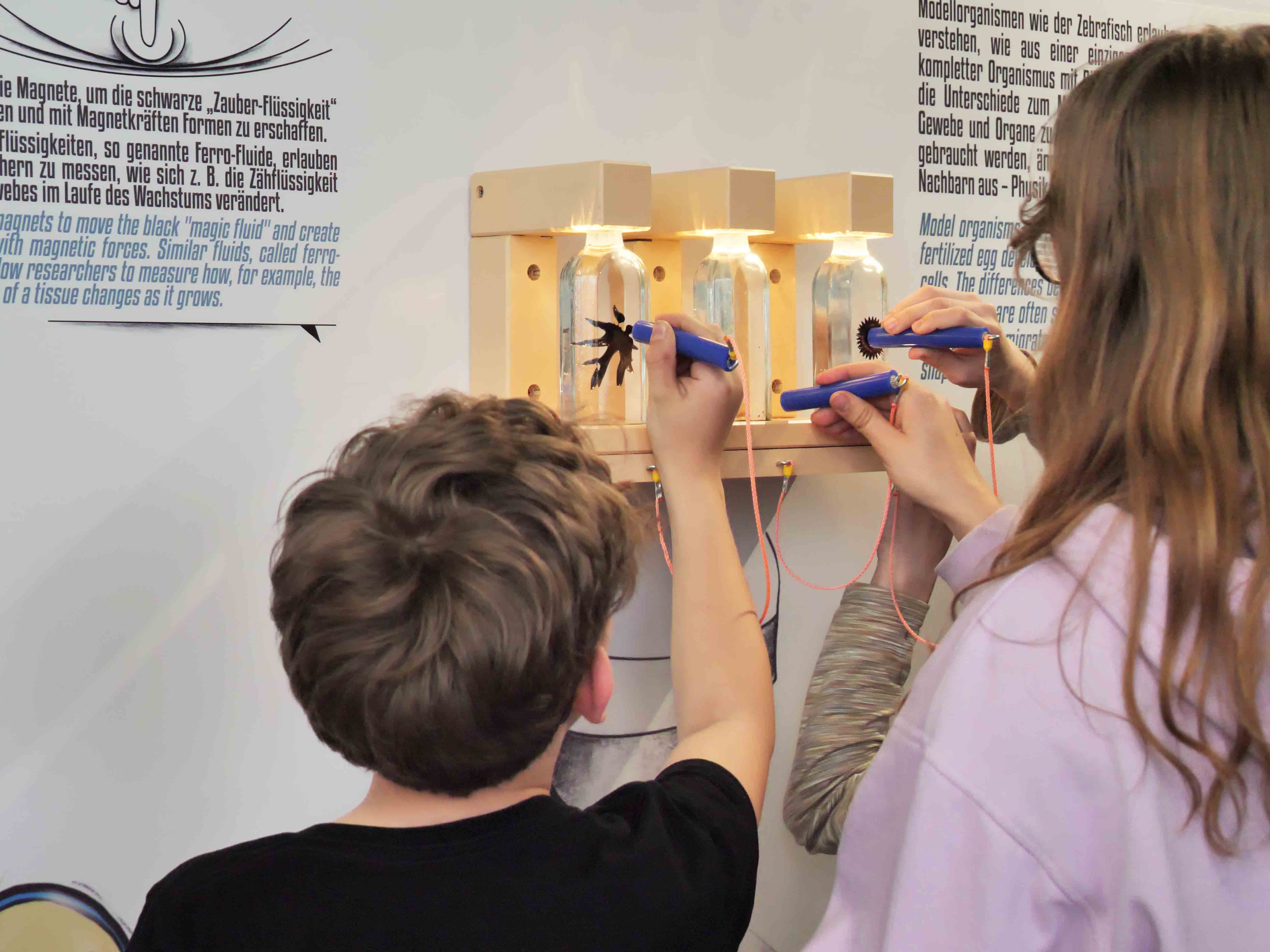 KI generiert: Das Bild zeigt zwei Kinder, die mit magnetischen Werkzeugen in Glasbehältern mit Ferrofluid experimentieren. Der Hauptinhalt des Bildes ist eine interaktive Wissenschaftsaktivität zum Thema Magnetismus und Ferrofluid.