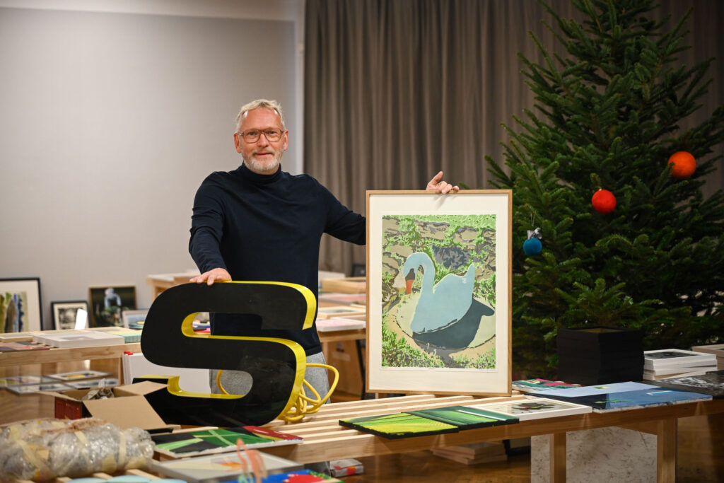 KI generiert: Ein Mann steht neben einem Tisch und hält ein gerahmtes Bild eines Schwans hoch. Neben ihm befindet sich ein Tannenbaum mit bunten Kugeln geschmückt.