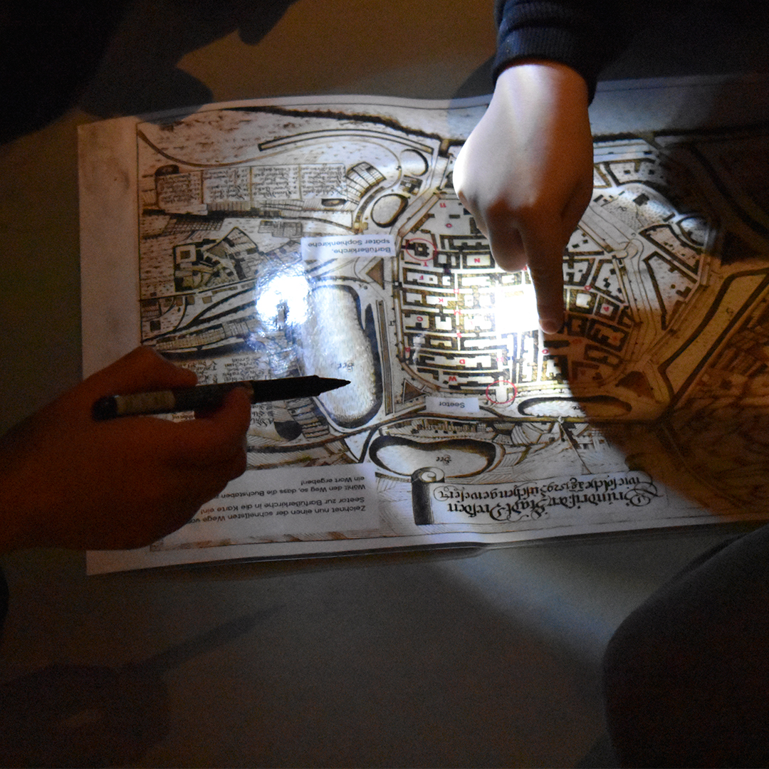 KI generiert: Das Bild zeigt zwei Hände, die auf einen alten Stadtplan zeigen, der von einem Lichtstrahl beleuchtet wird. Eine Hand hält einen Stift, während die andere auf einen bestimmten Punkt auf der Karte deutet.
