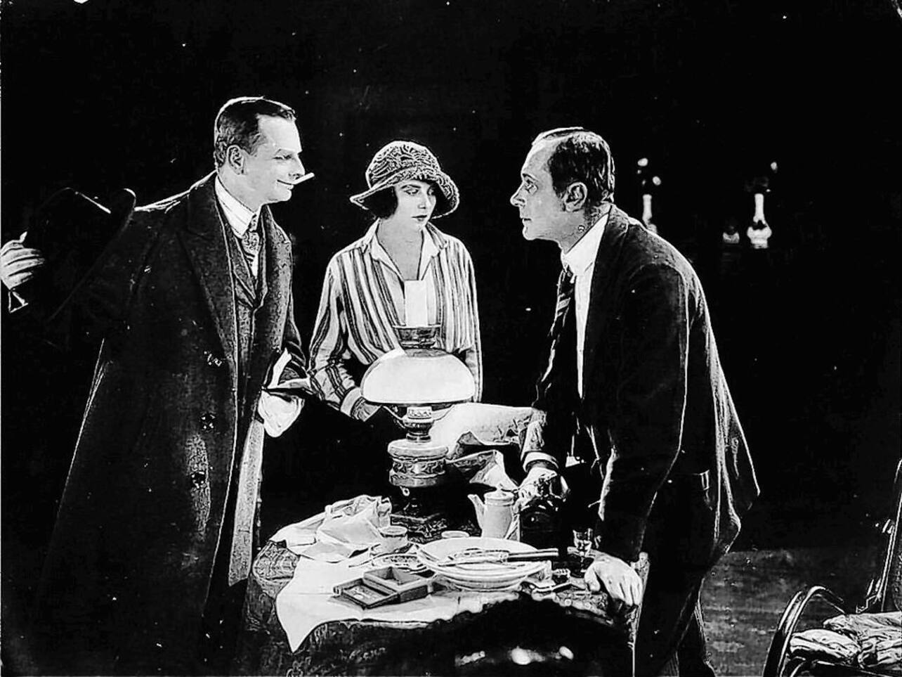 KI generiert: Das Bild zeigt drei Personen, die um einen Tisch mit einer Lampe in der Mitte versammelt sind, wobei zwei Männer sich angeregt unterhalten und die Frau in der Mitte steht und zusieht. Die Szene wirkt dramatisch mit einem Fokus auf Interaktion und Spannung zwischen den Charakteren.