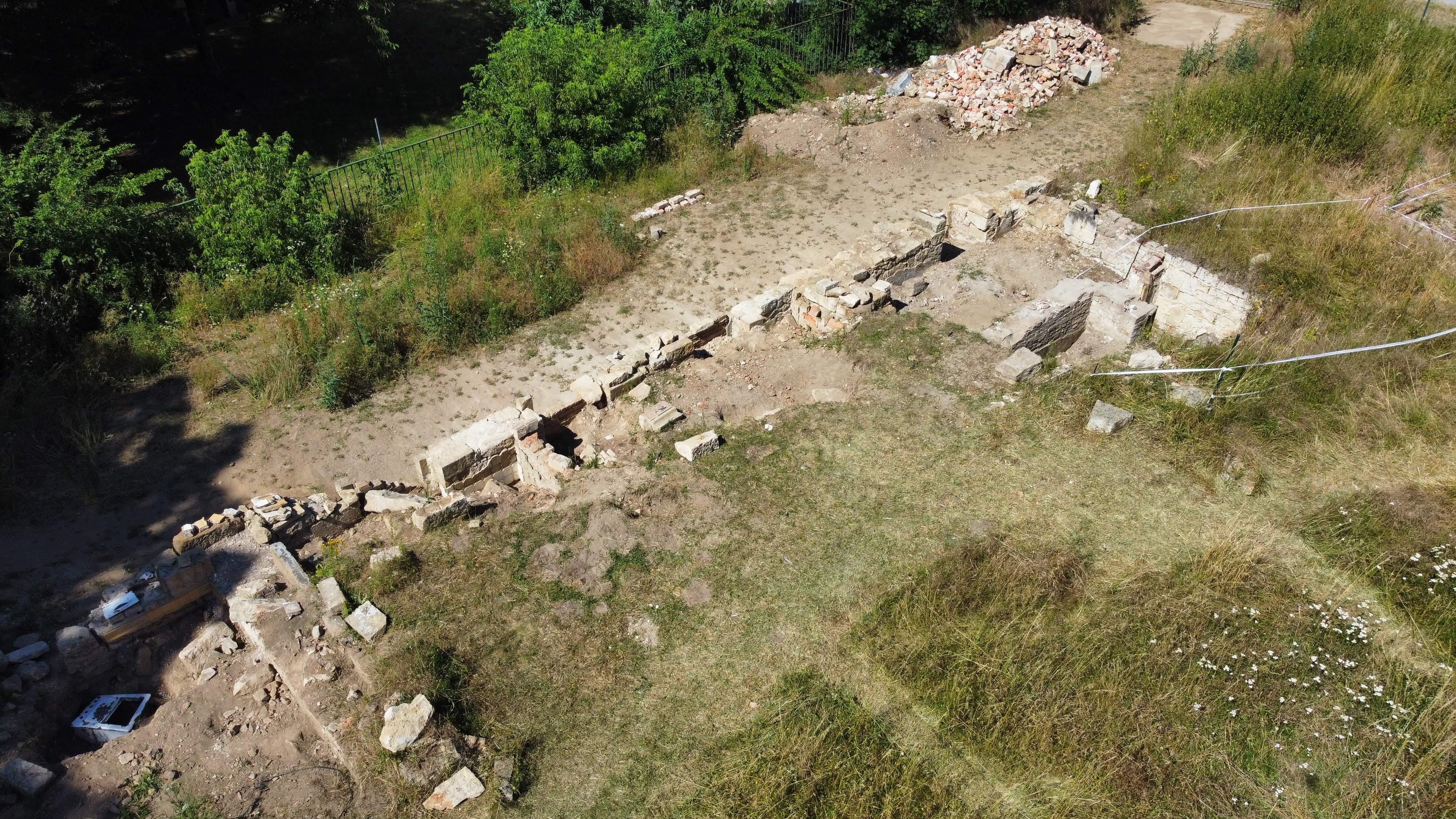 KI generiert: Das Bild zeigt eine archäologische Ausgrabungsstätte mit freigelegten Mauerresten und umgebender Vegetation. Im Hintergrund sind aufgestapelte Steine oder Trümmer zu sehen.