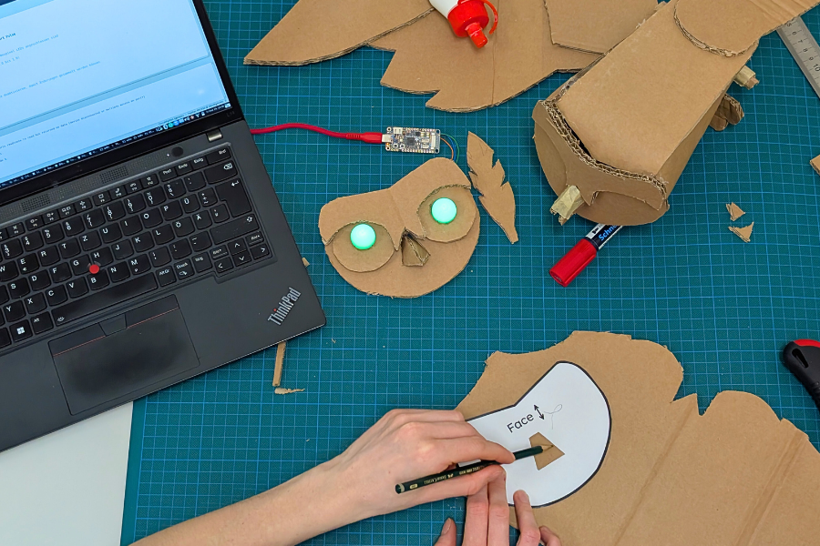 KI generiert: Ein Bastelprojekt ist im Gange, bei dem jemand aus Pappe einen Eulenkopf mit leuchtenden Augen erstellt und ein Laptop zur Unterstützung nutzt. Auf dem Tisch liegen verschiedene Werkzeuge und Materialien verstreut.