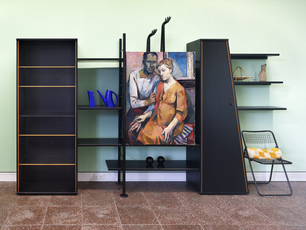 AI generated: The image shows a modern shelving and furniture system in a room with a light green wall. A painting depicting a seated person and a standing person hangs in the centre.