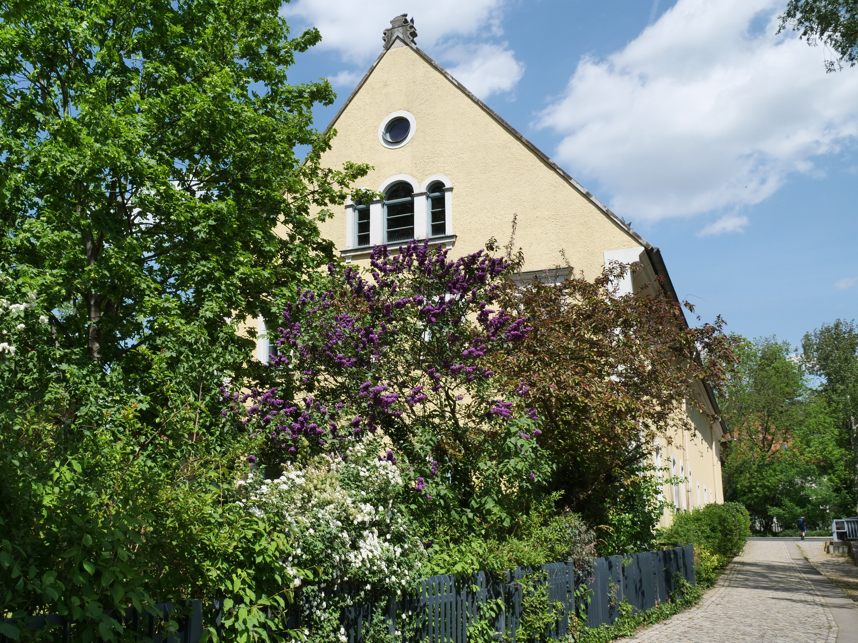 KI generiert: Das Bild zeigt ein gelbes, traditionelles Wohnhaus mit einem roten Ziegeldach und Garten, im Hintergrund steht ein großes modernes Hochhaus. Im Vordergrund ist eine von Bäumen und Grünflächen umgebene Treppe zu sehen.