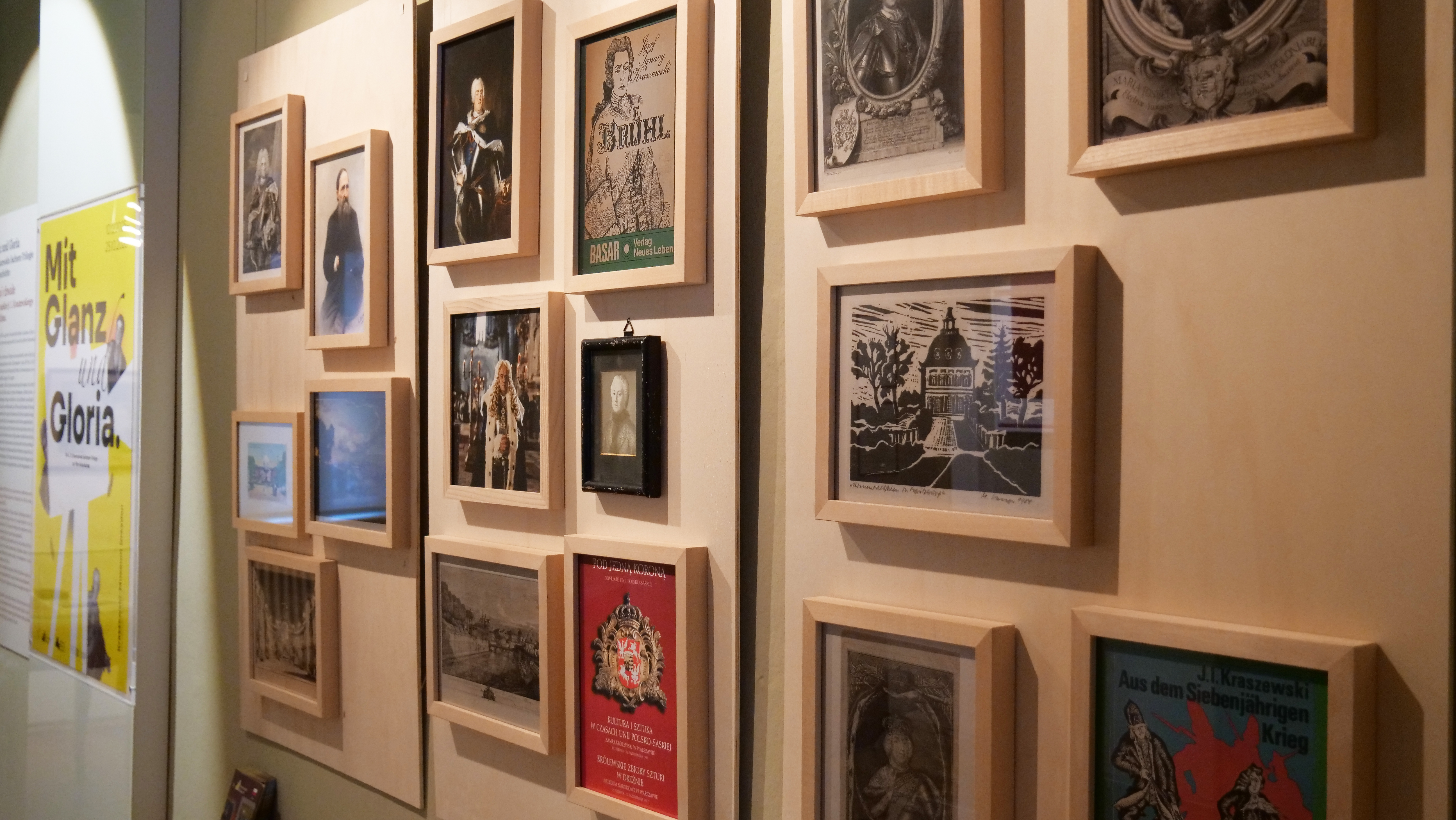 KI generiert: Das Bild zeigt eine Wand mit einer Sammlung von gerahmten Bildern und Plakaten, die wahrscheinlich historische oder künstlerische Themen darstellen. Links ist ein gelbes Plakat mit der Aufschrift "Mit Glanz & Gloria" zu sehen.