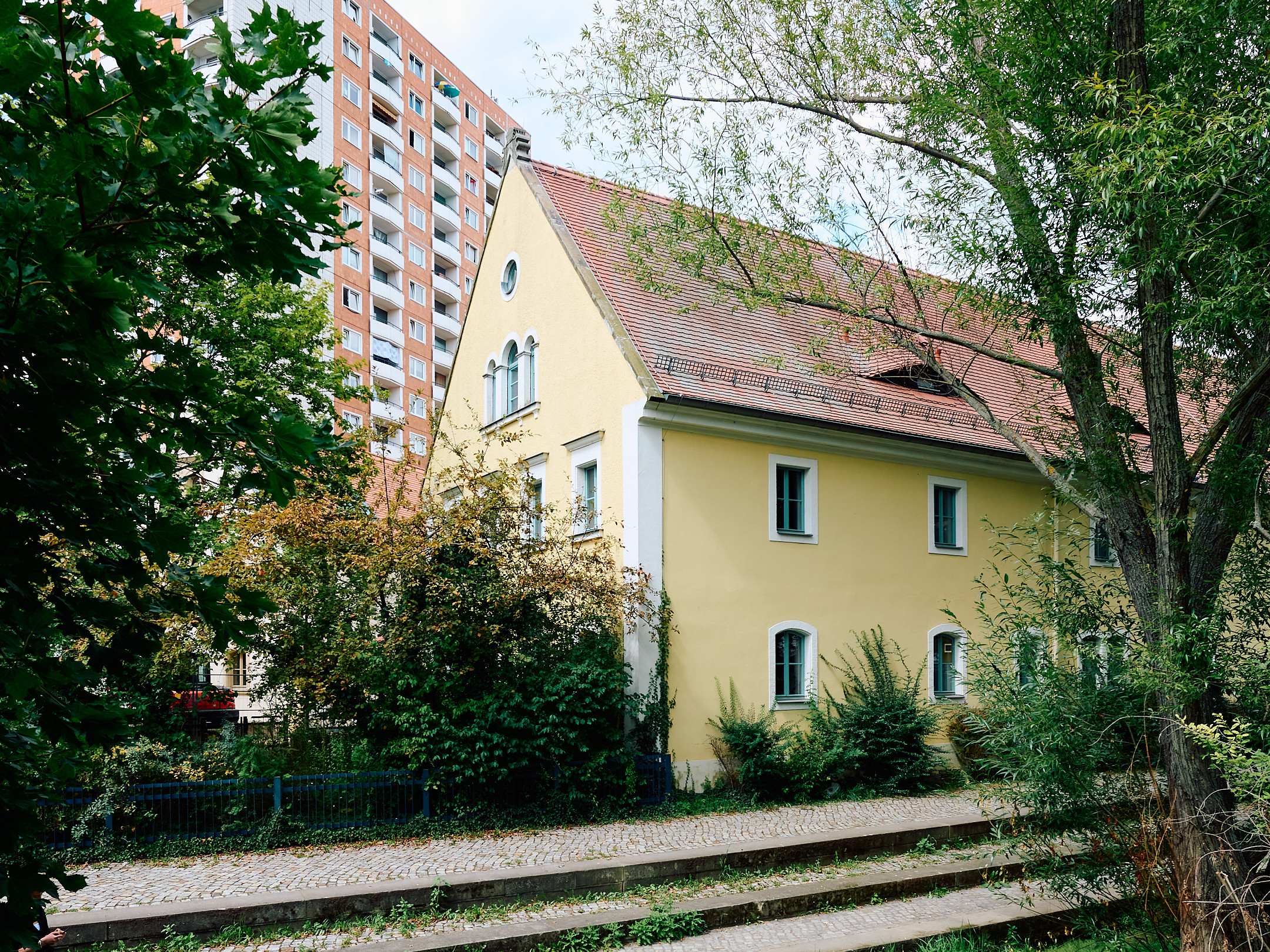 KI generiert: Das Bild zeigt ein gelbes, traditionelles Wohnhaus mit einem roten Ziegeldach und Garten, im Hintergrund steht ein großes modernes Hochhaus. Im Vordergrund ist eine von Bäumen und Grünflächen umgebene Treppe zu sehen.