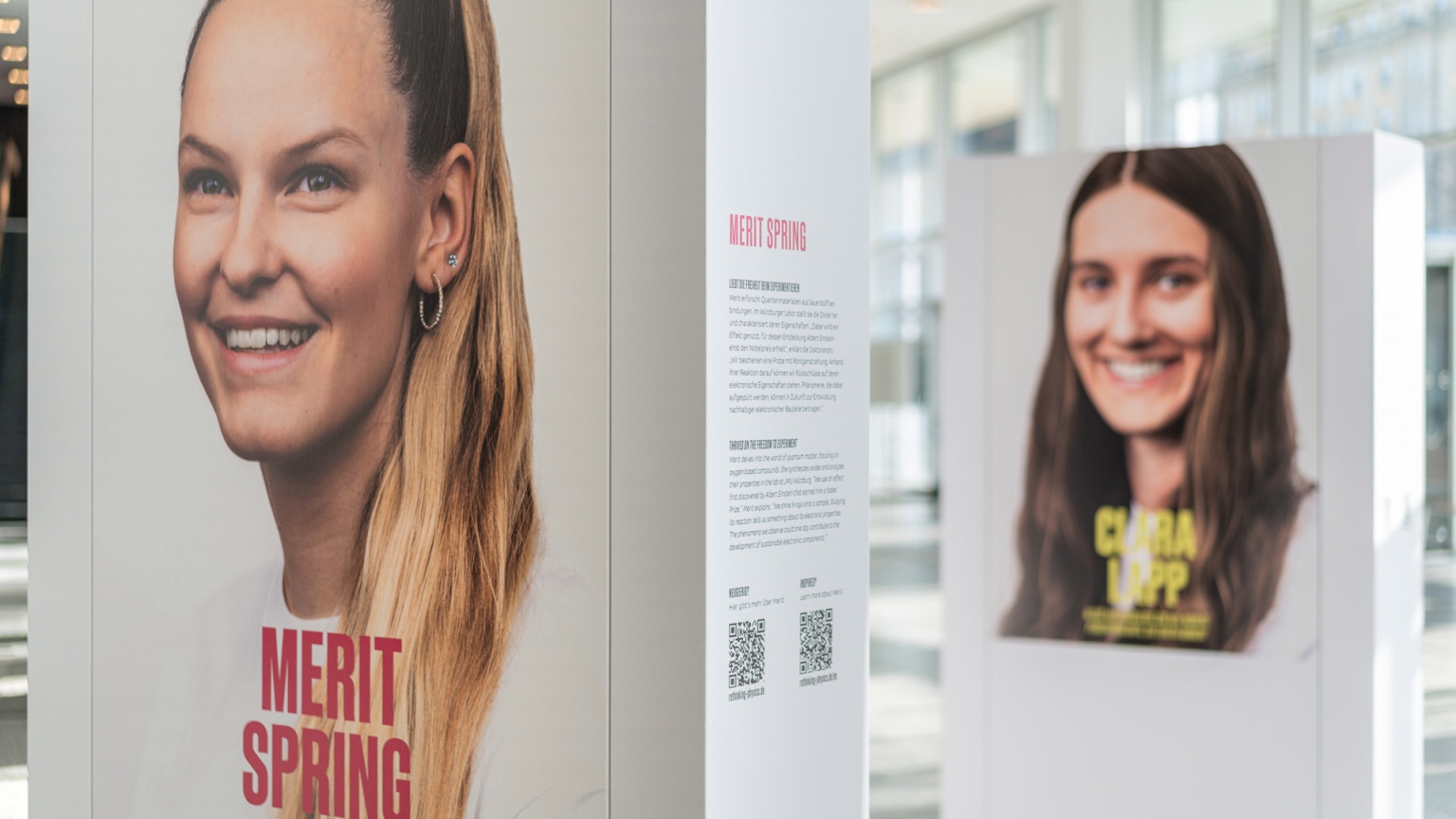 KI generiert: Das Bild zeigt eine Ausstellung mit großformatigen Porträtfotos und Texten, die sich auf Ausstellungstafeln befinden. Im Vordergrund ist ein Porträt einer lächelnden Frau mit dem Namen "Merit Spring" zu sehen.