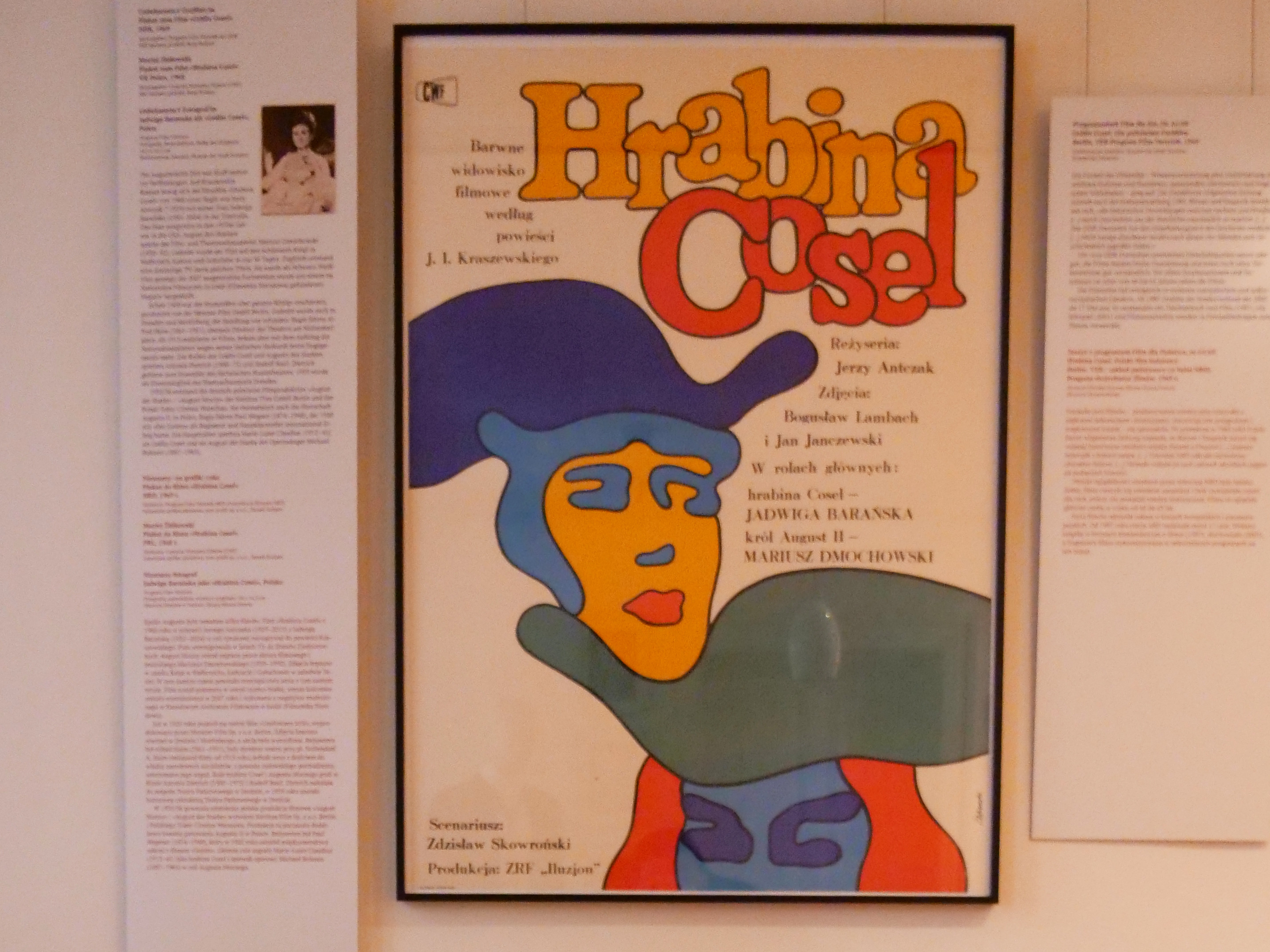 KI generiert: Das Bild zeigt im Hintergrund ein buntes Plakat mit dem Titel "Hrabina Cosel" an einer Wand, flankiert von zwei Texttafeln. Im Vordergrund liegt eine Zeitschrift mit dem Titel "NBI" auf einem Tisch.