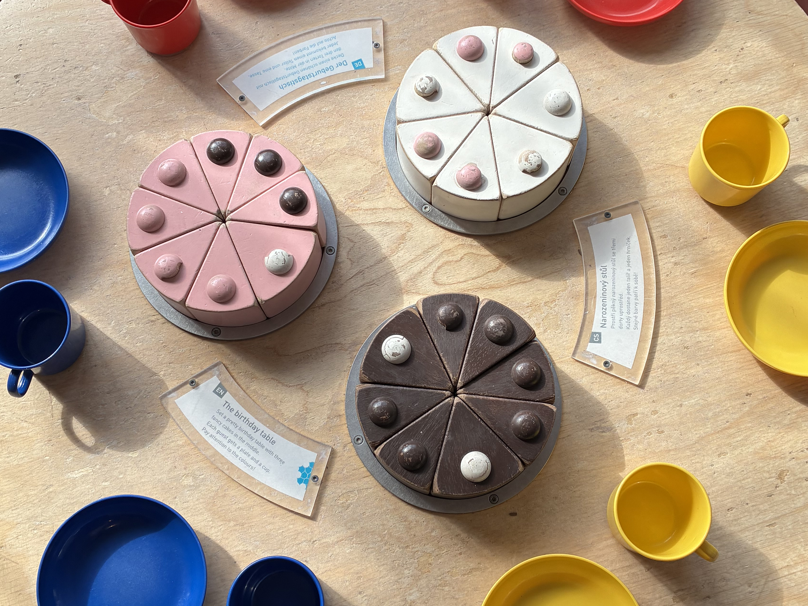 KI generiert: Auf dem Bild sind drei runde, in Segmente unterteilte Kuchenattrappen in verschiedenen Farben (rosa, weiß und braun) sowie bunte Tassen und Teller auf einem Tisch zu sehen. Daneben liegen Schilder mit Text, die vermutlich die Modelle oder Szenarien beschreiben.